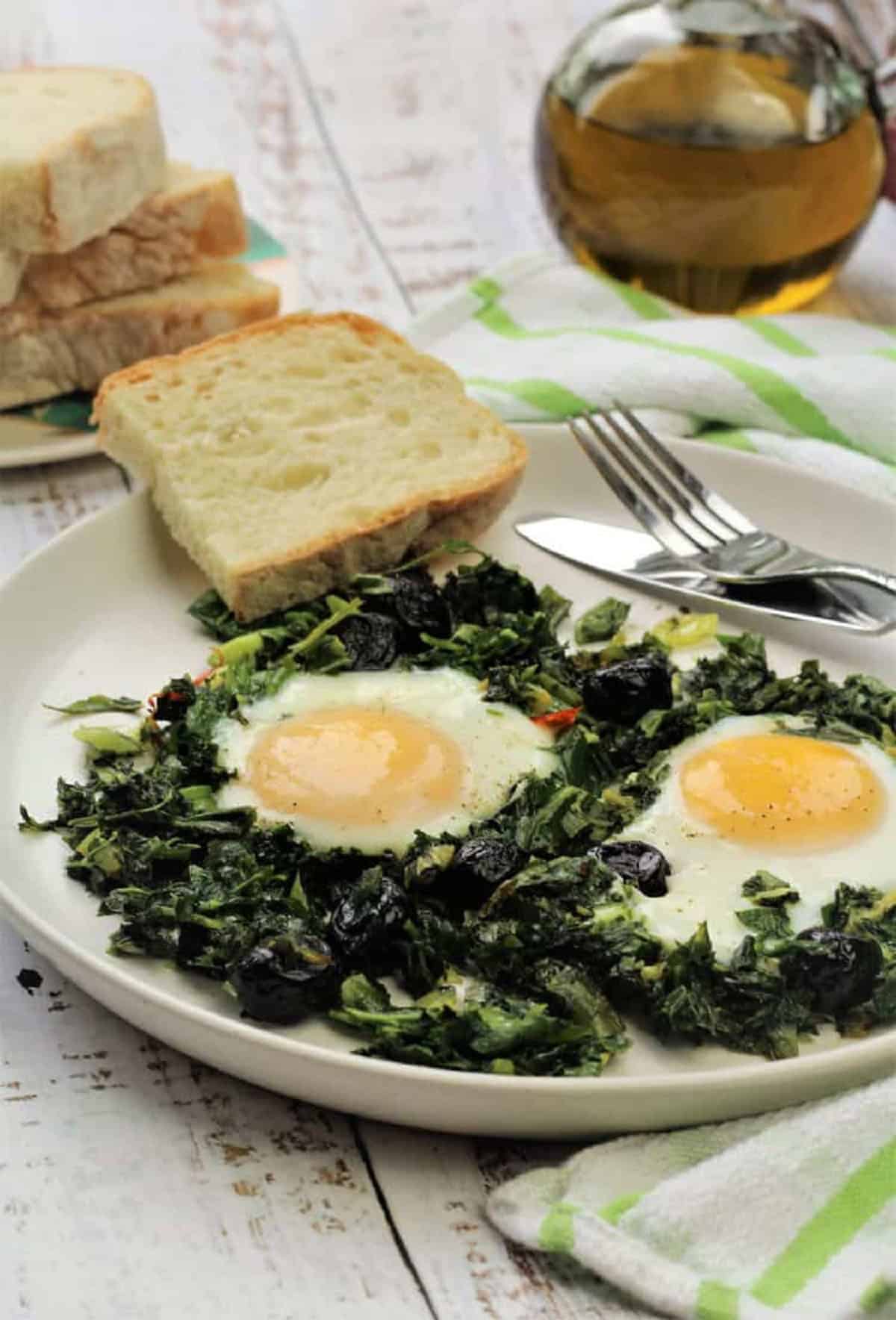 Two fried eggs with greens and black olives on plate with sliced bread.