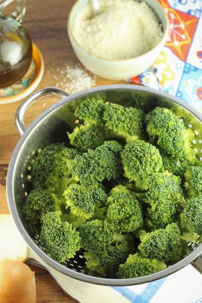 Broccoli divided into florets in colander next to olive oil bottle and bowl of grated Parmigiano cheese.