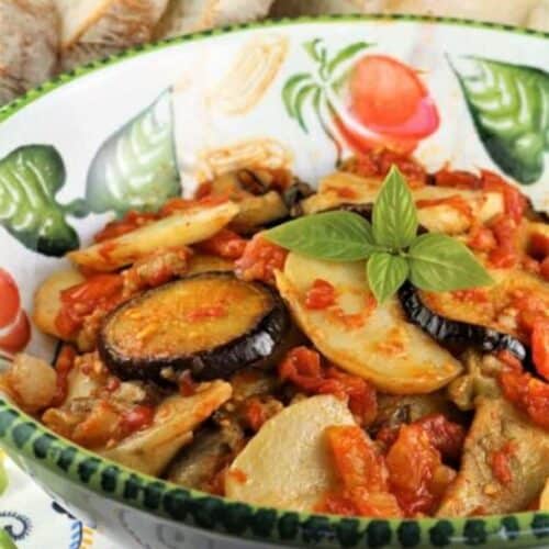 Baked eggplant and potatoes with tomato sauce in large serving bowl topped with fresh basil.