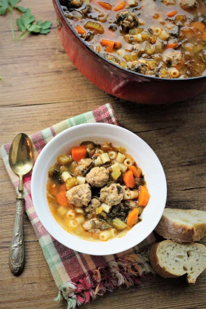 red pot with vegetable and meatball soup with bowl of soup, spoon and sliced bread next to it