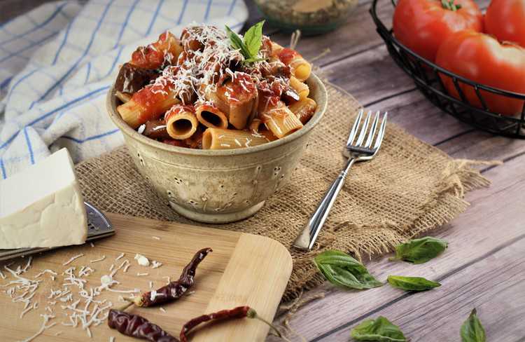 ceramic bowl filled with pasta and eggplant in sauce next to cheese wedge, basil leaves and chili peppers