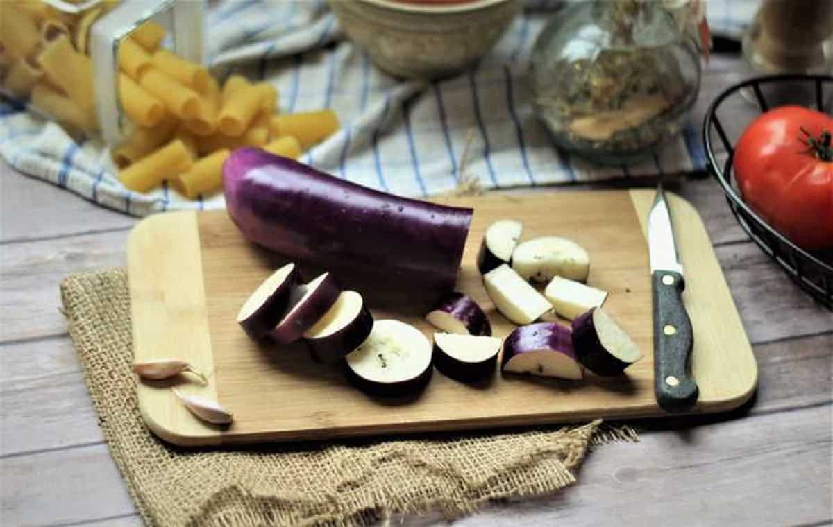 Sliced eggplant on wood board surrounded by tomatoes, olive oil and rigatoni pasta.