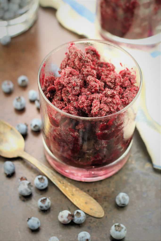 glass filled with blueberry granita with spoon and fresh blueberries around it