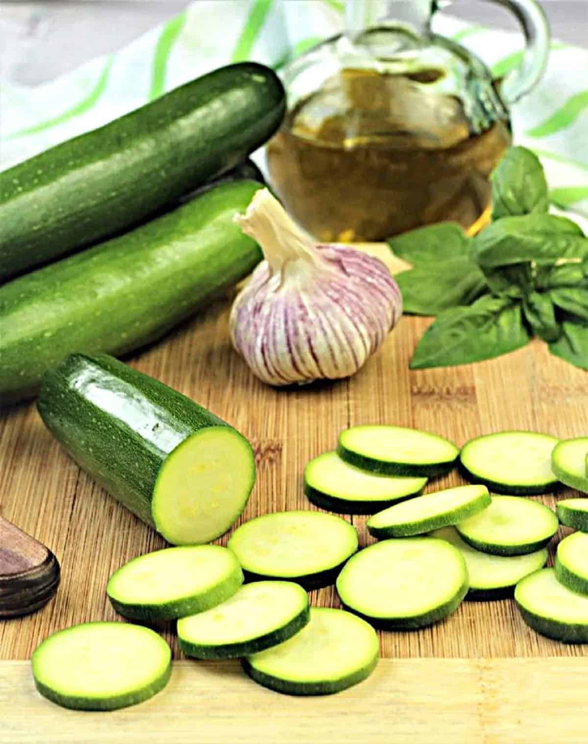 Sliced zucchini rounds on wood board with garlic bulb, basil and olive oil bottle.