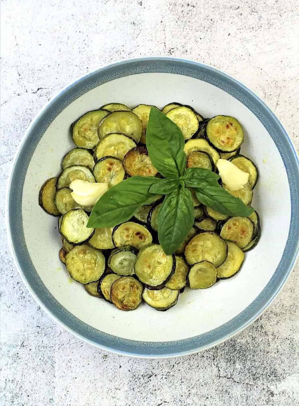 Roasted zucchini rounds in bowl with clove of garlic and basil leaves.