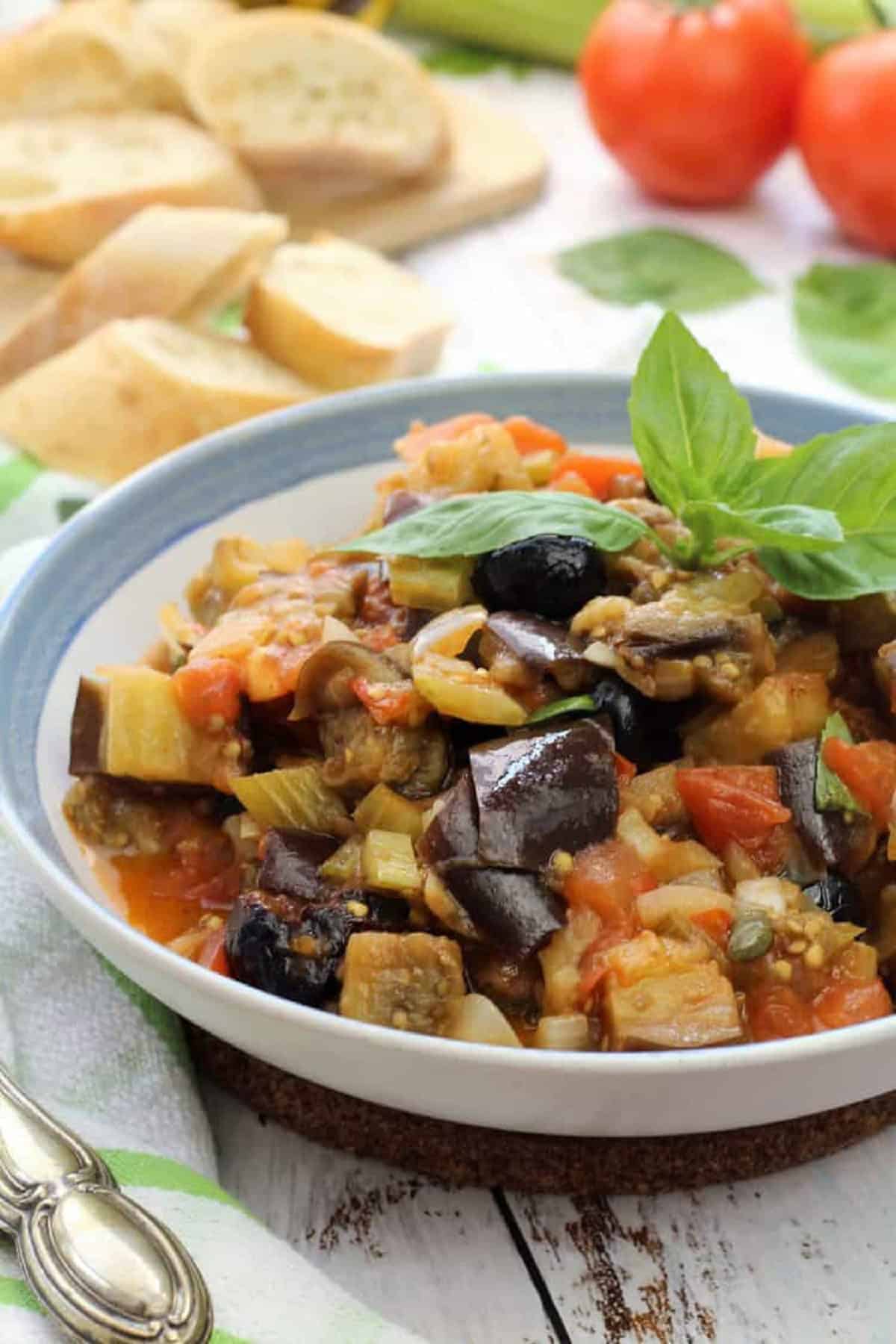 Sicilian eggplant caponata topped with fresh basil in bowl next to sliced bread and tomatoes.