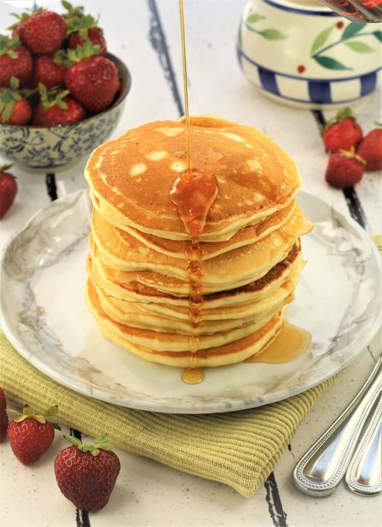 stack of yogurt pancakes with maple syrup drizzled on top and bowl of strawberries in background