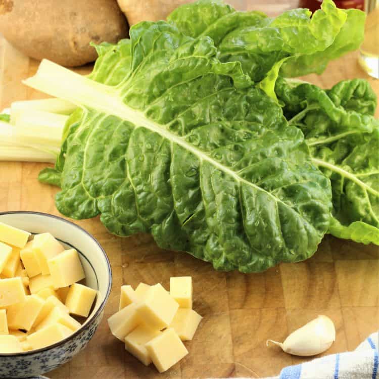 swiss chard leaves on cutting board with bowl of cubed cheese and whole potatoes in background