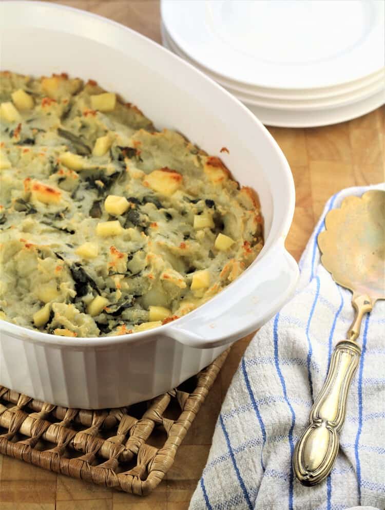 white casserole with potato casserole with serving spoon on side and stack of plates behind it