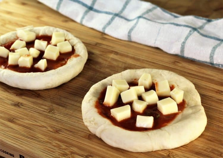 two unbaked mini pizzas with tomato sauce and cubed mozzarella on wooden board