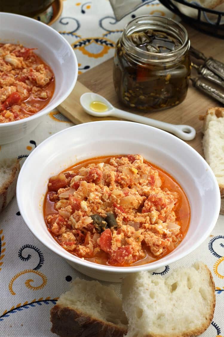 bowl of fried tomatoes with eggs alongside bread and jar of chili peppers