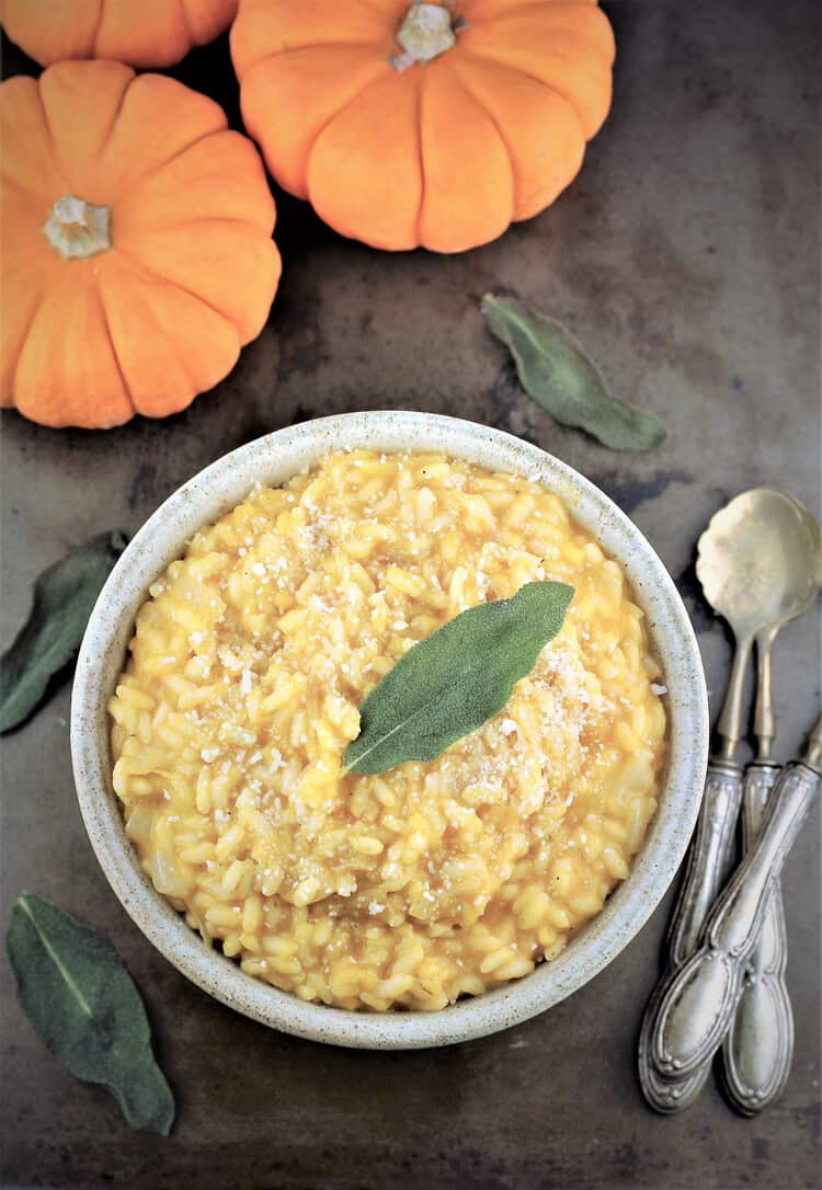 bowl of pumpkin risotto with crispy sage leaf on top and mini pumpkins and spoons on side