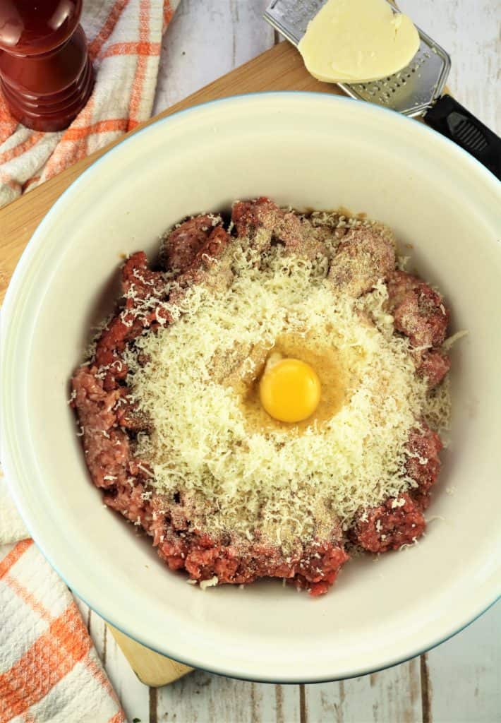 large mixing bowl with ground meat, cheese, breadcrumbs and egg with cheese on grater and pepper mill