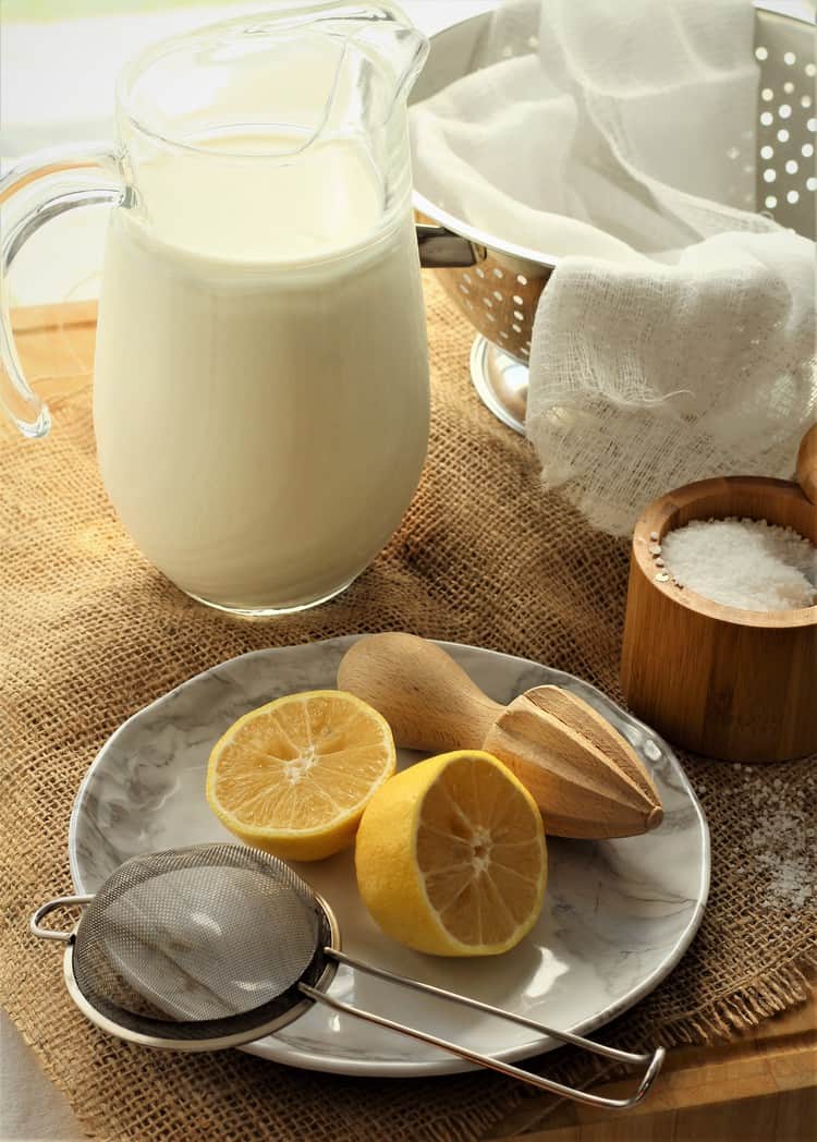 pitcher of milk, colander lined with cheese cloth and halved lemon with juicer and sieve on plate