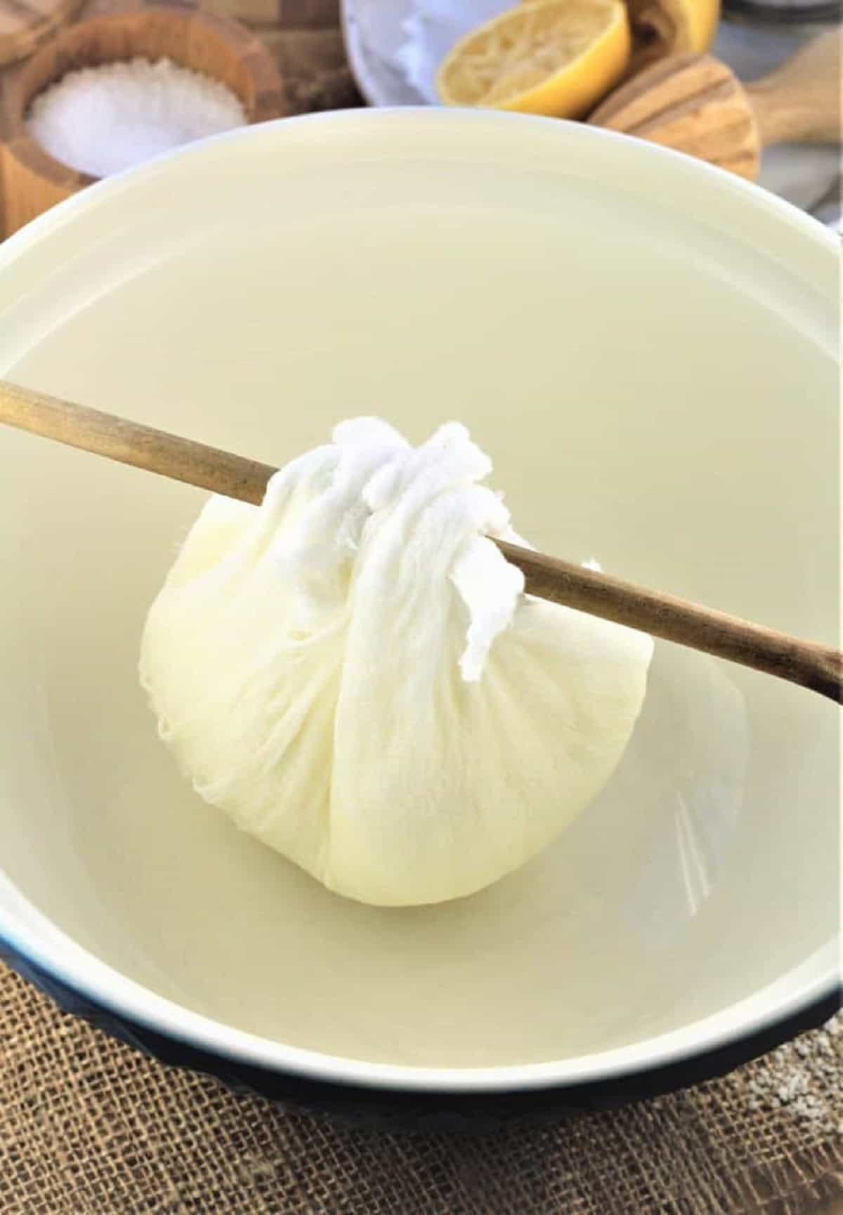 Ricotta in cheese cloth tied on a wooden spoon over a bowl to drain excess liquid.