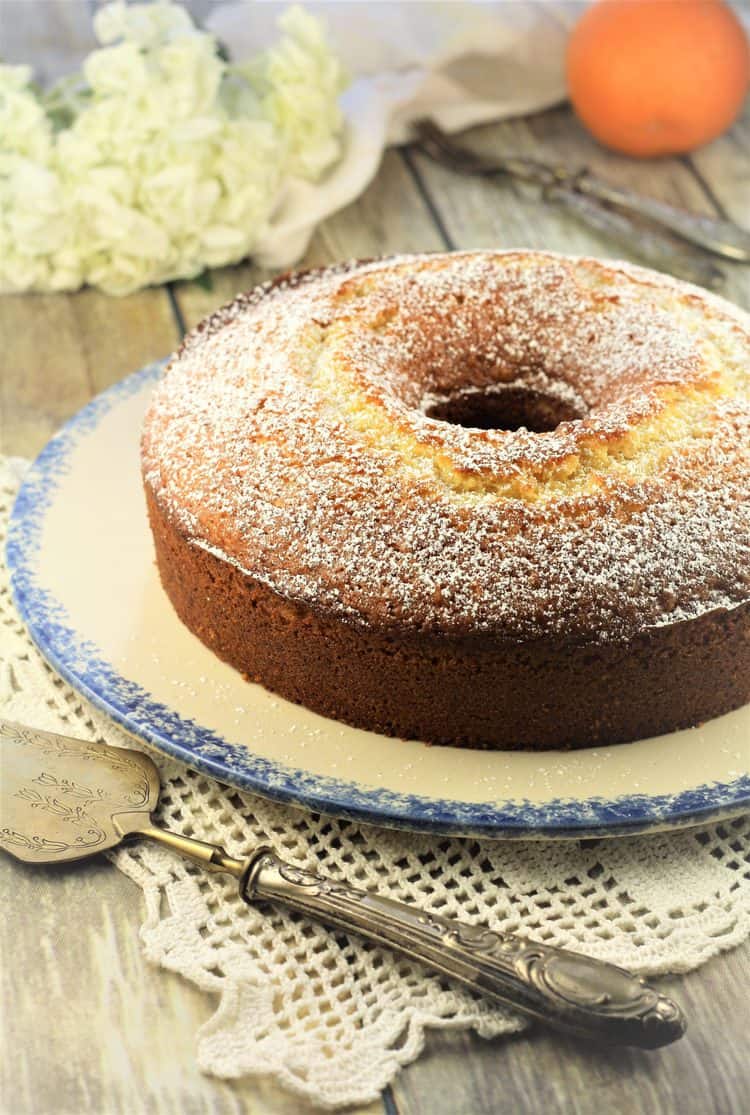 bundt cake on blue rimmed plate with cake spatula and flowers and orange in background