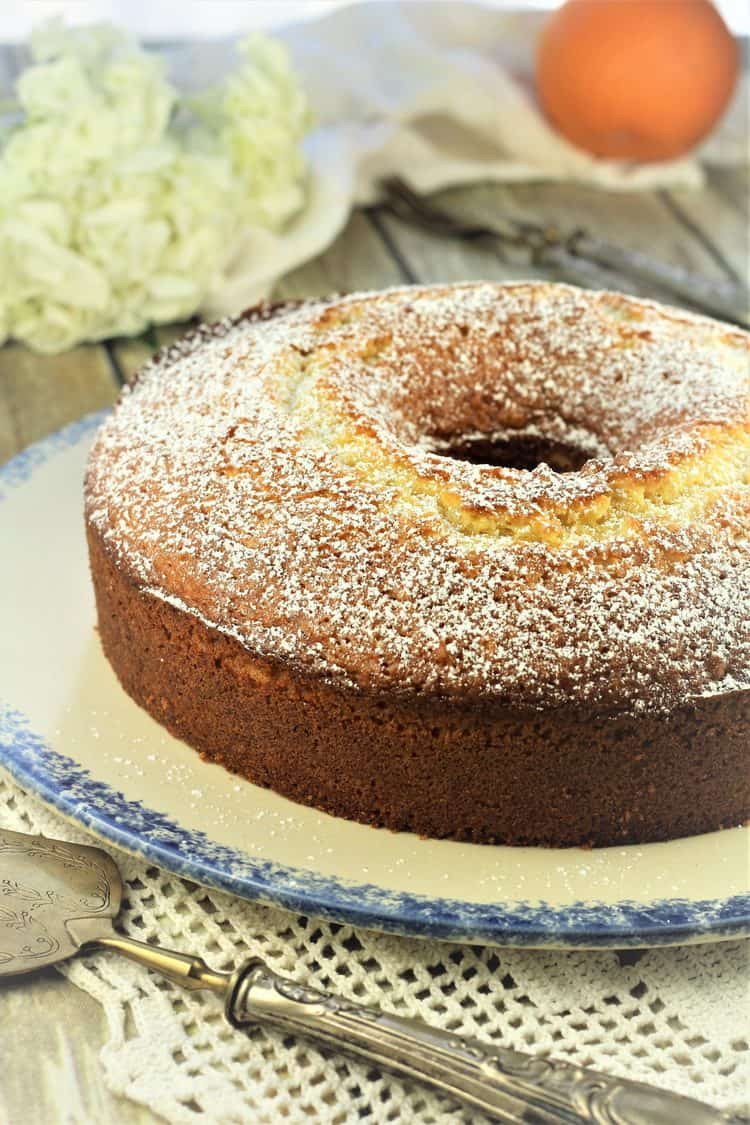 orange bundt cake on blue rimmed plate with cake spatula and flowers and orange in background