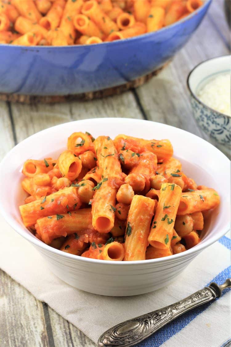 white bowl filled with pasta with chickpeas in tomato sauce with blue skillet filled with pasta and bowl of cheese behind it