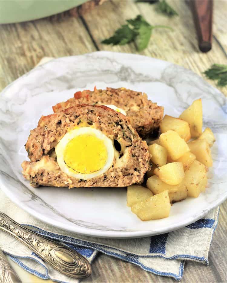 closeup of polpettone with hard boiled eggs and roast potatoes on white plate with utensils on side