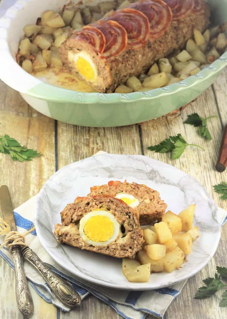 sliced polpettone with hard boiled eggs on plate surrounded by roast potatoes and whole meatloaf in roasting pan behind it