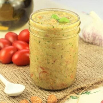 Sicilian pesto alla Trapanese in glass jar surrounded by almonds, cherry tomatoes, garlic bulb and olive oil.