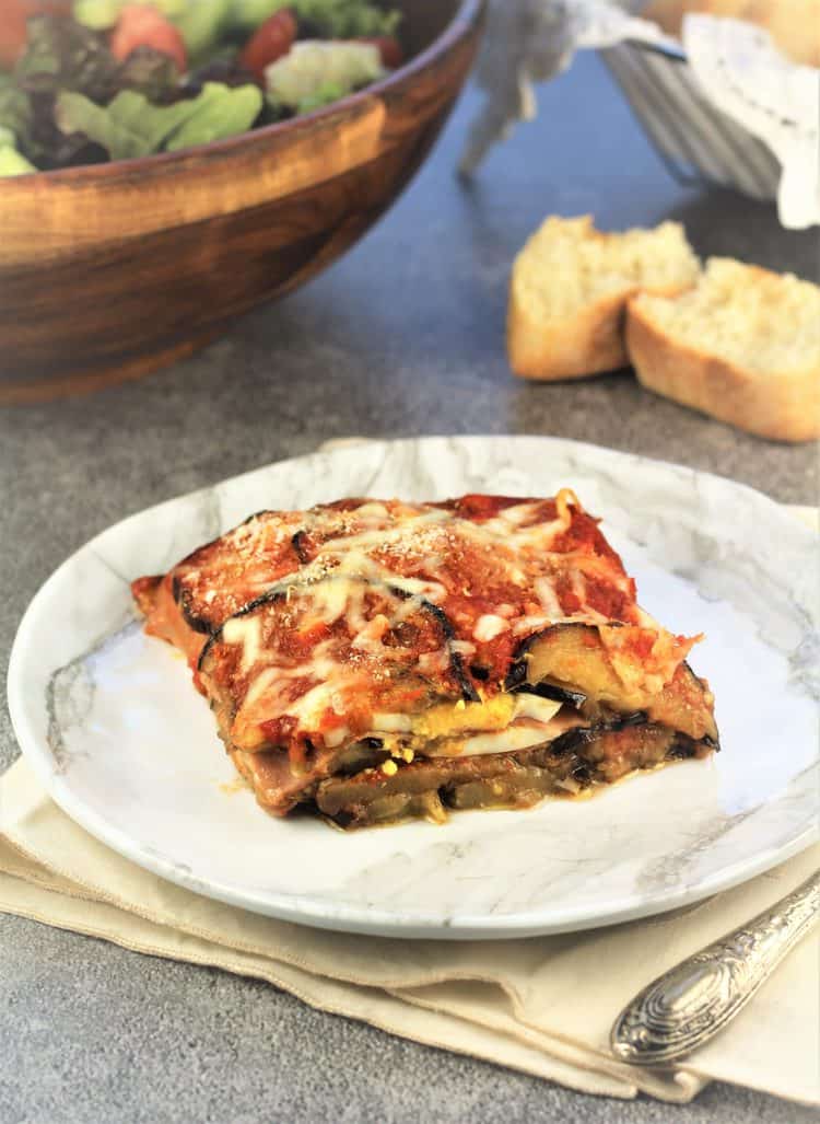 piece of eggplant parmigiana on white plate with bread slices and bowl of salad in background