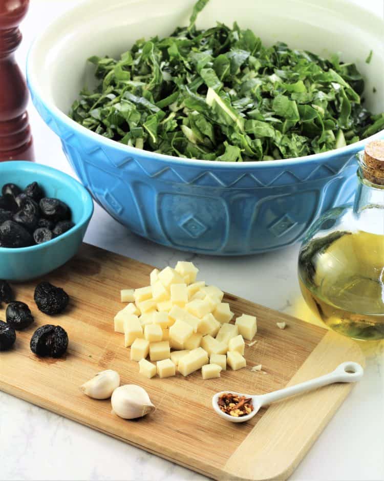 chopped swiss chard in large blue bowl with cubed cheese, black olives and olive oil flask in forefront