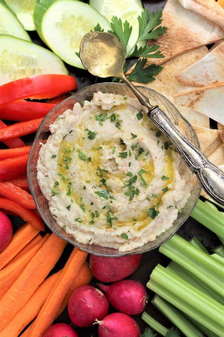 white bean dip in glass bowl with spoon over it surrounded by crudités