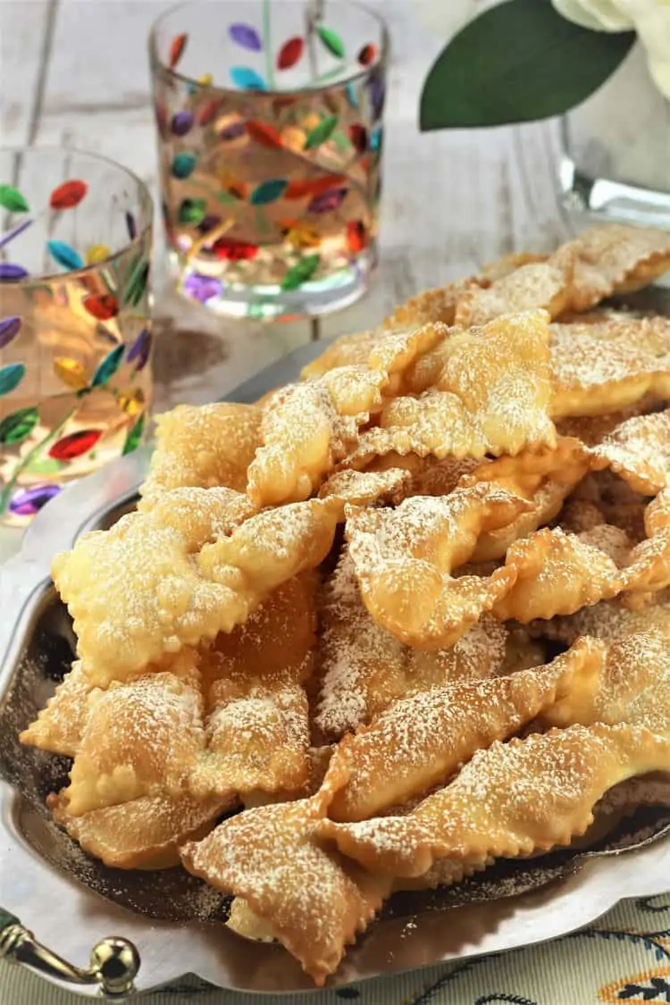 pile of chiacchiere on silver tray with 2 colorful goblets next to it