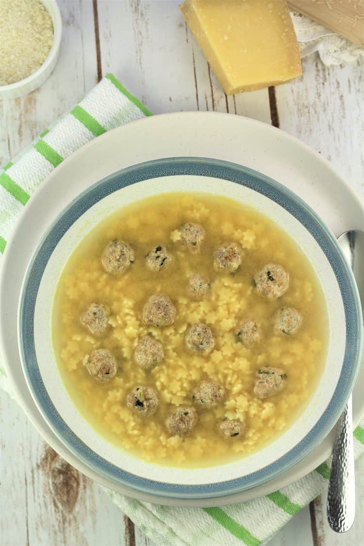 blue rimmed bowl filled with pastina soup with tiny meatballs with spoon on side and cheese in background