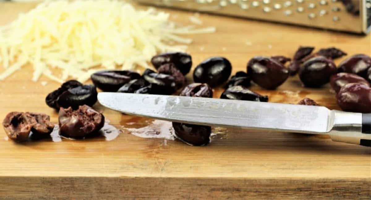 Pitting black olives with the flat side of a knife on wood board next to shredded cheese.
