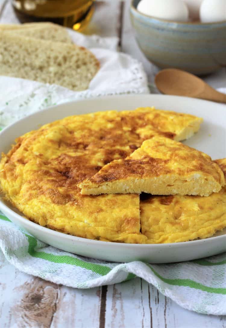 white plate with ricotta frittata cut into wedges with sliced bread and bowl of eggs behind it