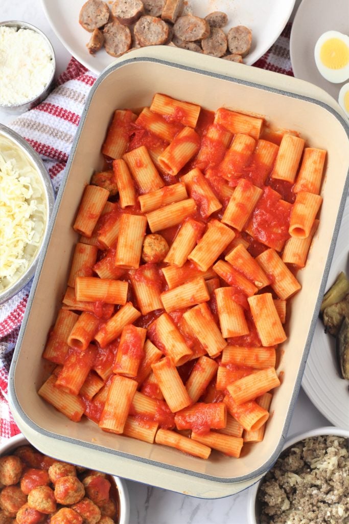 rigatoni with tomato sauce in baking dish surrounded by bowls with ingredients for pasta al forno