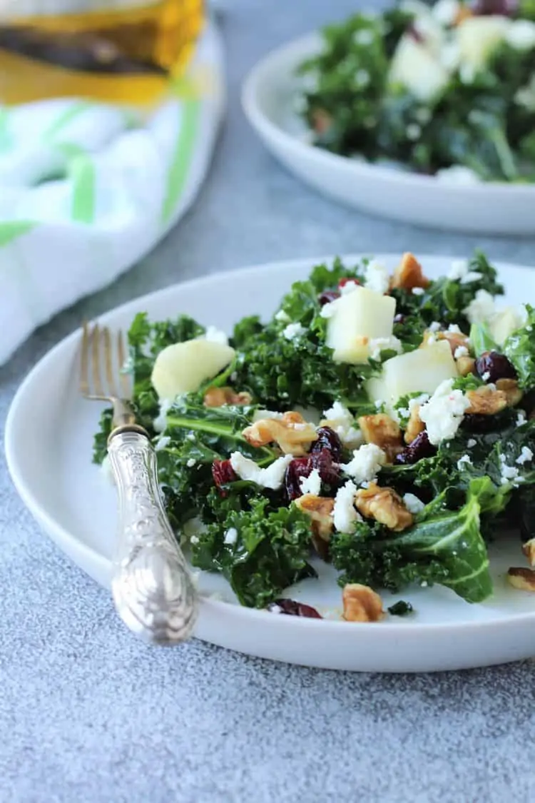 white plate with kale salad topped with apples, walnuts, cranberries and feta with fork on side