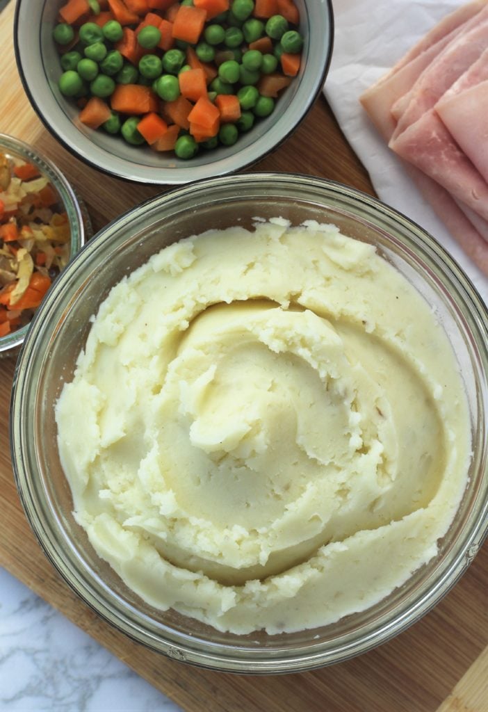 glass bowl with mashed potatoes, bowl of carrots and peas on wood board 