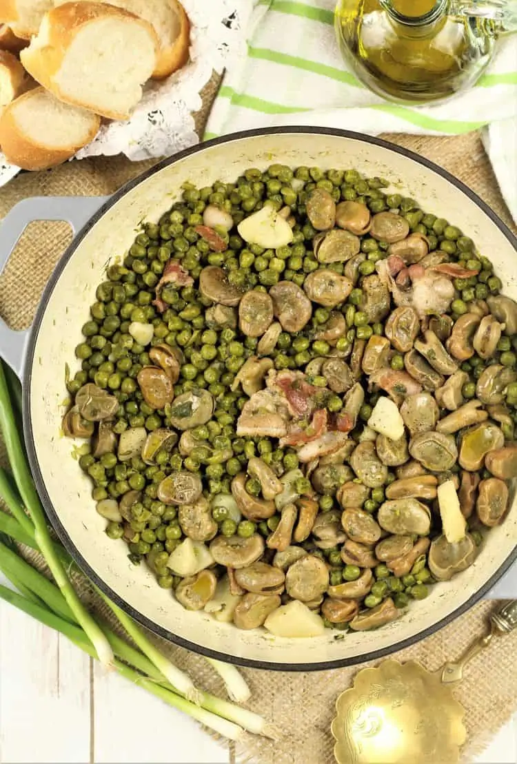 large skillet filled with fava bean and pea stew with green onions, sliced bread and olive oil bottle on side
