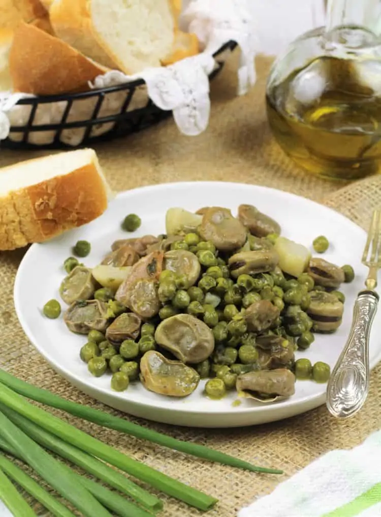 white plate filled with fava beans and peas with wedge of bread, green onions and olive oil bottle around it