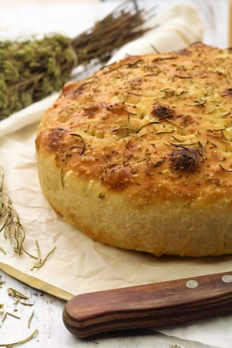 round herbed focaccia bread on wood board with knife and dry herbs 