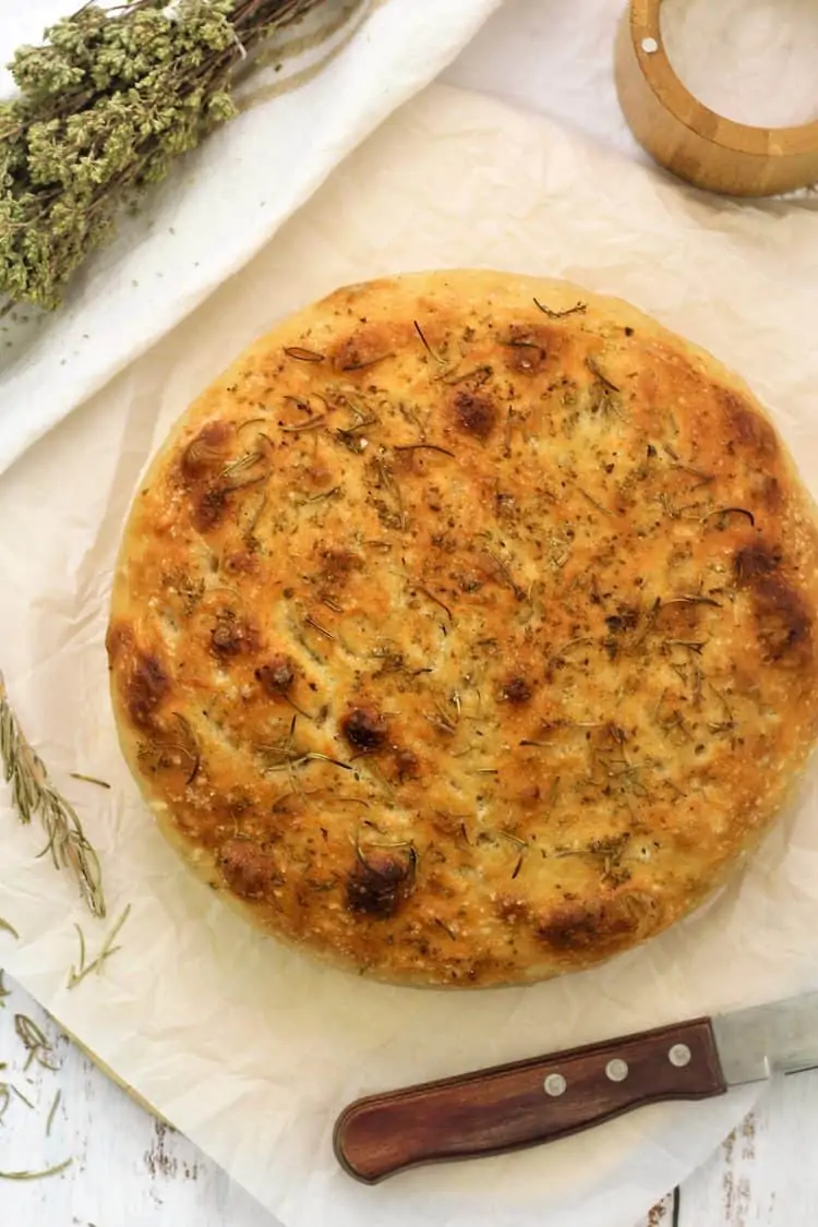 round herbed focaccia on parchment paper surrounded by herbs