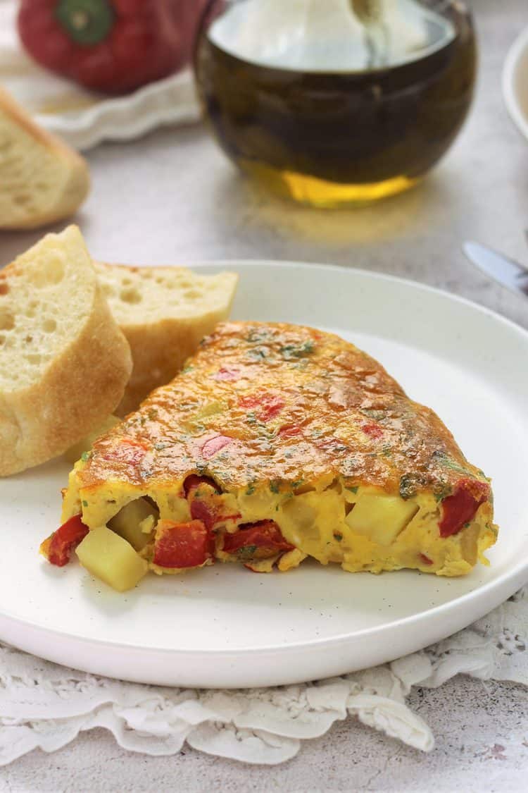 potato and red pepper frittata wedge on round white plate with sliced bread and olive oil bottle behind it