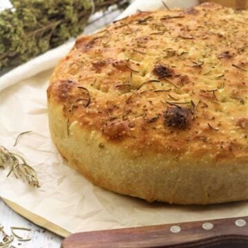 No knead skillet focaccia bread topped with herbs on wood board with knife and oregano branches.