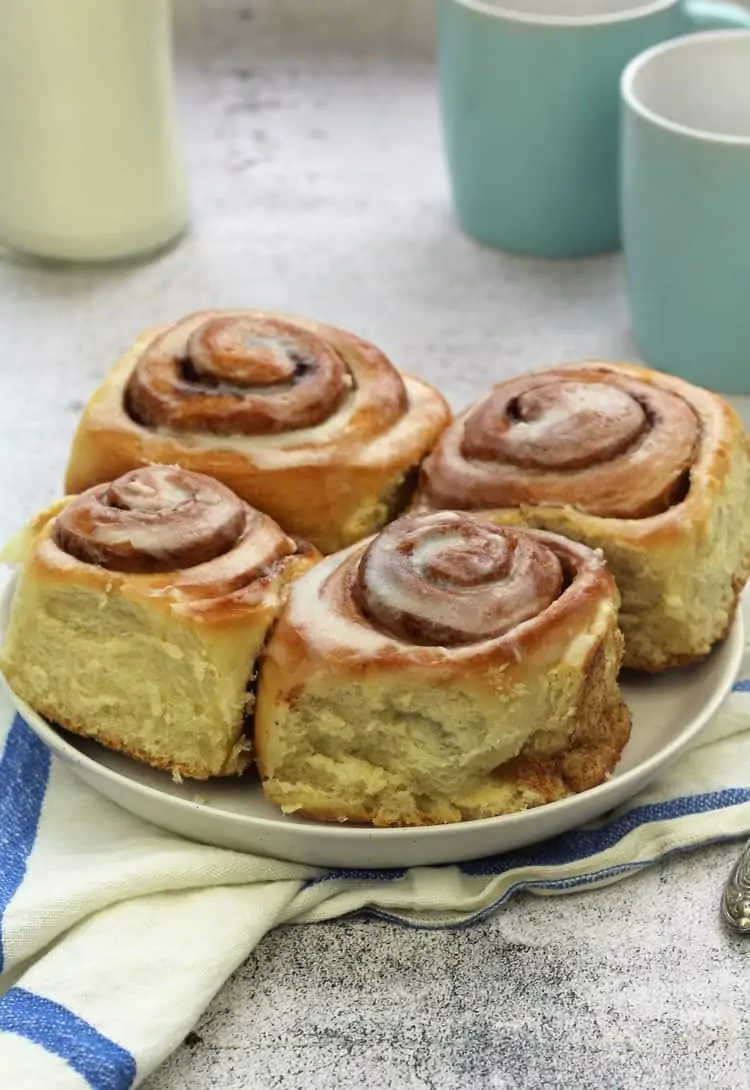 cinnamon rolls on round white plate with blue coffee mugs and bottle of milk