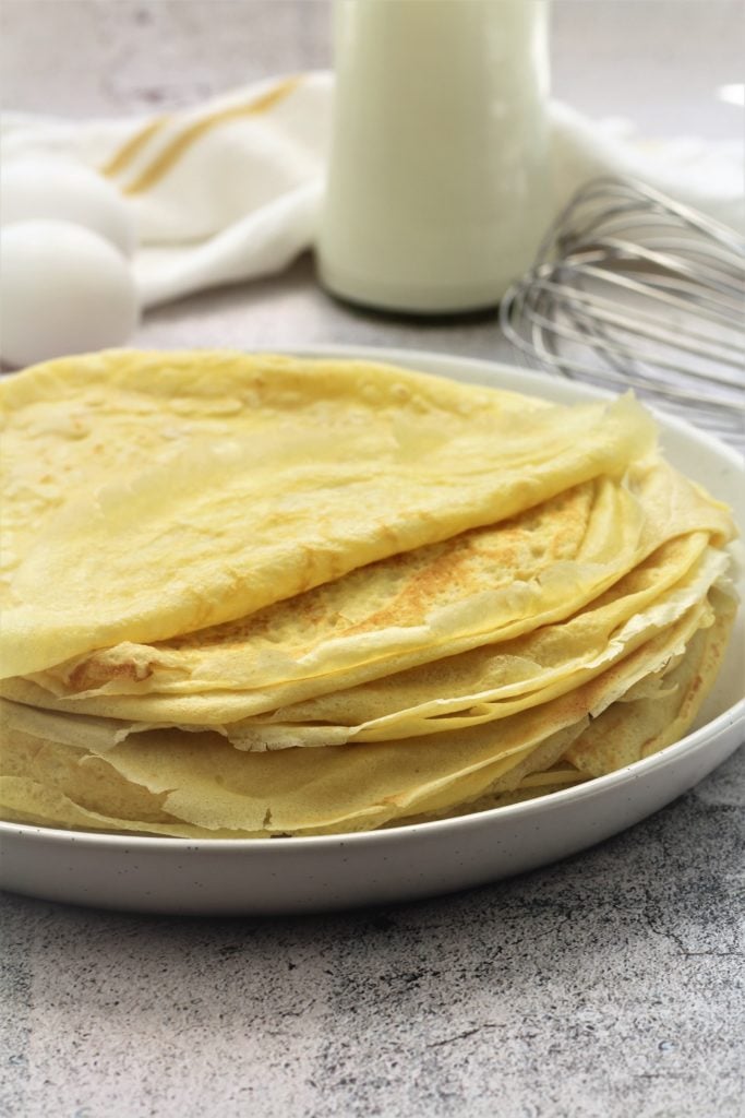 stack of crepes on round plate with eggs, milk bottle and whisk in background