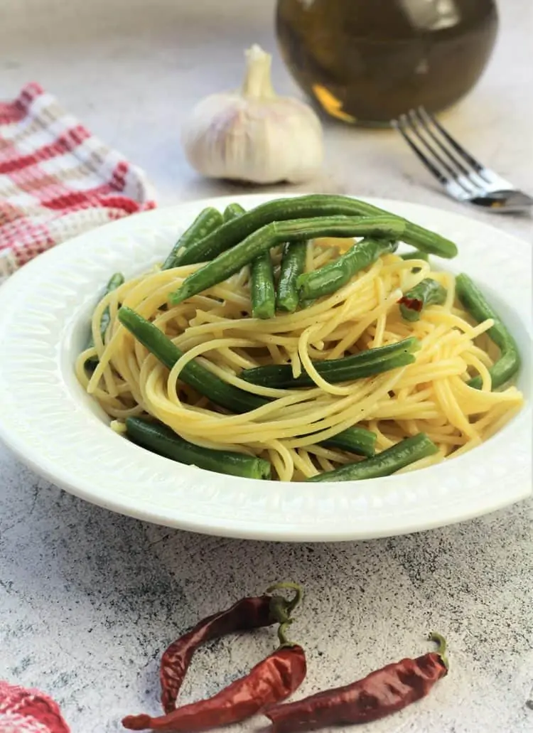 white bowl filled with spaghetti with green beans surrounded by garlic bulb, olive oil, fork and red chilis