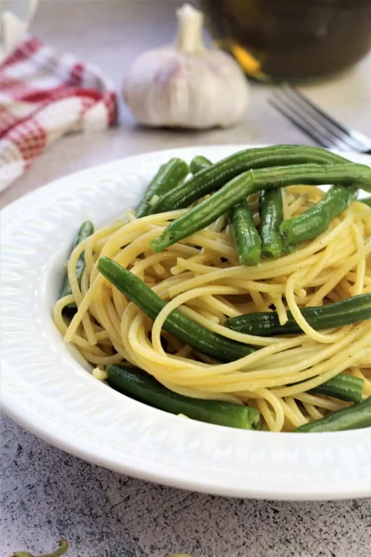 white bowl filled with spaghetti with green beans with garlic bulb, fork and olive oil behind it