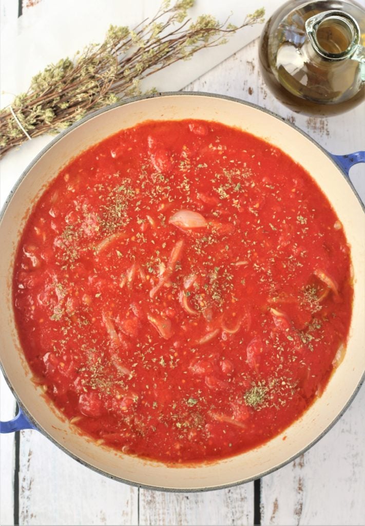 large skillet filled with simmering tomato sauce with oregano branches and olive oil bottle on side 