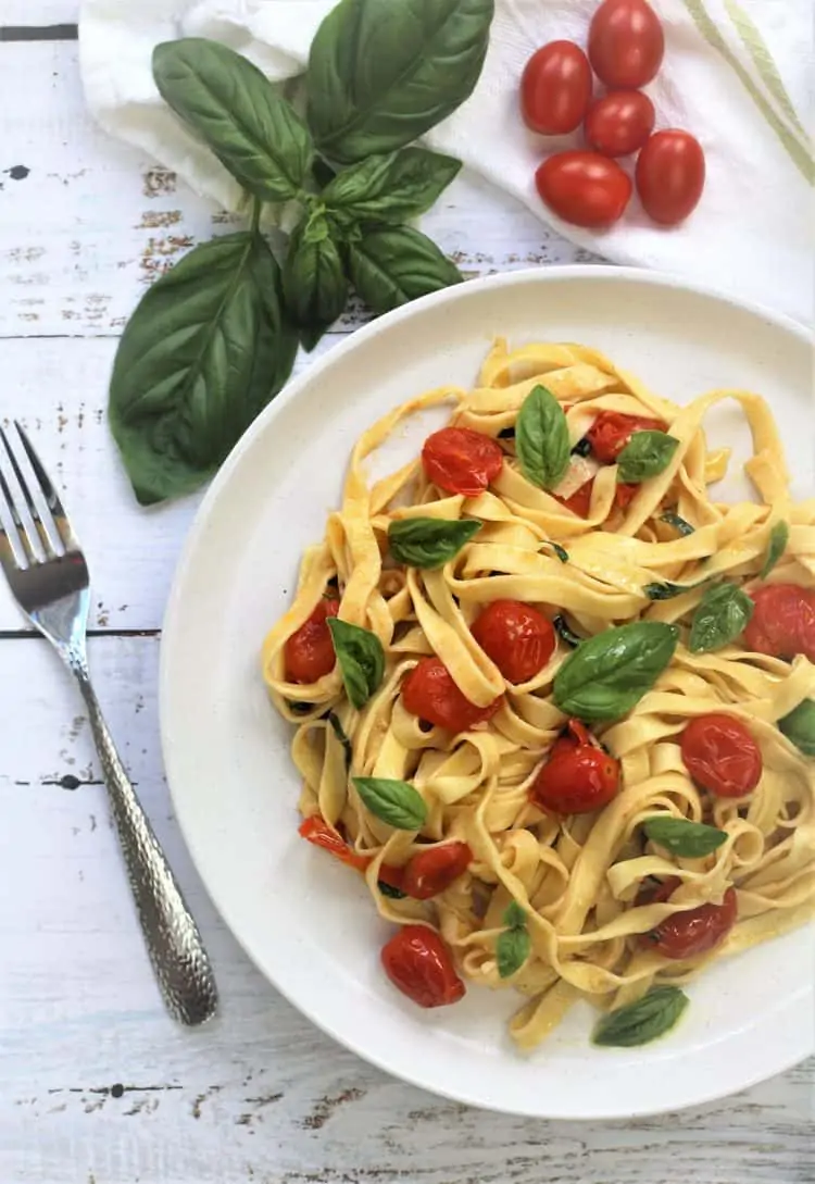 white plate filled with Homemade Tagliatelle with Cherry Tomatoes, Garlic and Basil with fork on side and basil leaves and cherry tomatoes behind it