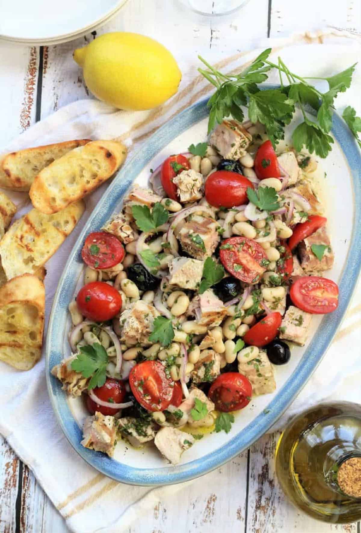 Tuna ,white bean and tomato salad on oval plate with crostini, lemon and olive oil bottle.