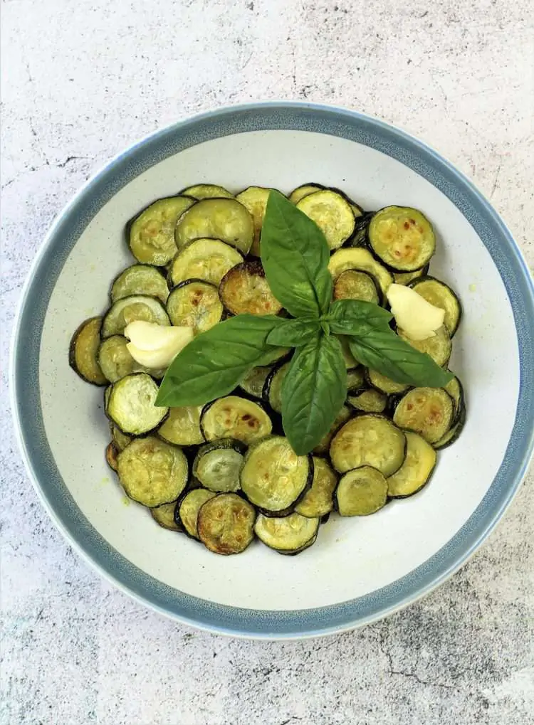 roasted zucchini slices in blue rimmed bowl topped with basil leaves and garlic clove