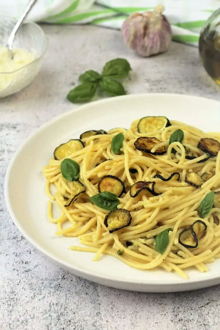 white plate with spaghetti with oven fried zucchini surrounded by cheese bowl, basil leaves and garlic bulb